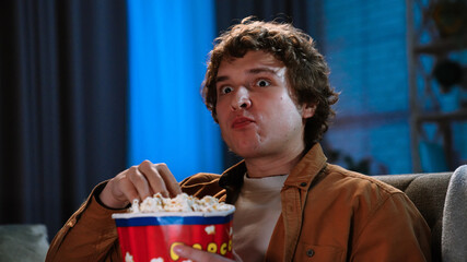 Young man spending evening at home sitting on sofa in living room, watching movie on tv, eating popcorn from basket, amazed face expression, close up.