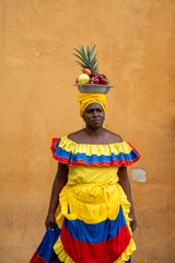 A Palenquera Woman Wearing A Red, Yellow, And Blue Dress, Holding Out Her Skirt With A Fruit Basket Balanced On Her Head