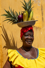 Woman In A Colorful Colombian Dress Balancing A Bowl Of Tropical Fruits Against A Yellow Wall