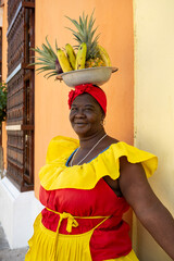 Colombian Woman In Red And Yellow Dress Balancing A Bowl Of Fruits In Front Of A Yellow Wall