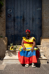A Palenquera Woman Sitting On Stone Steps Wearing A Bright Dress, With A Fruit Basket Next To Her Against A Blue Door
