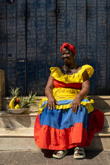 A Palenquera Woman Sitting On Stone Steps Wearing A Bright Dress, With A Fruit Basket Next To Her Against A Blue Door