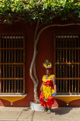 Woman In A Colorful Dress With A Fruit Basket On Her Head Standing Outside A Colonial Building With Wooden Windows