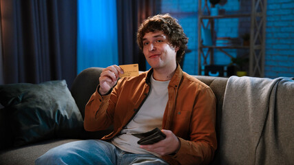 Fototapeta premium Young man spending evening at home sitting on sofa in living room, holding gold credit card in hand looking happy, smiling positive expression.