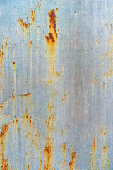 A close-up of a corroded metal surface with vertical streaks of orange and brown rust against faded blue-gray paint. The texture reveals weathered patterns, creating an aged, industrial appearance