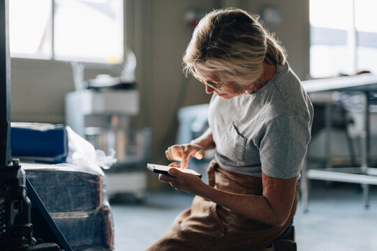 Senior female baker taking inventory of flour sacks through smart phone while working at bakery