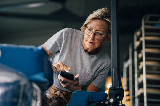 Low angle view of senior female baker taking inventory of flour sacks through smart phone while working at bakery