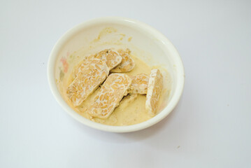 Raw tempeh that has been cut in a white bowl containing dough, with a white background.