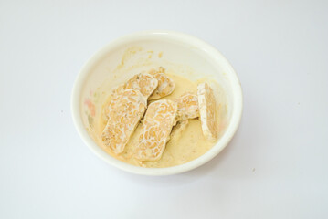 Raw tempeh that has been cut in a white bowl containing dough, with a white background.