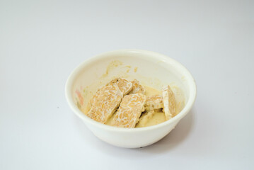 Raw tempeh that has been cut in a white bowl containing dough, with a white background.