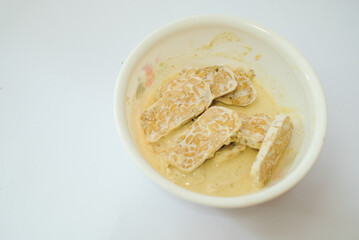 Raw tempeh that has been cut in a white bowl containing dough, with a white background.
