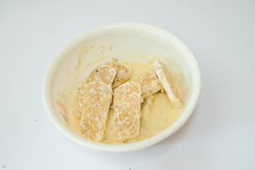 Raw tempeh that has been cut in a white bowl containing dough, with a white background.