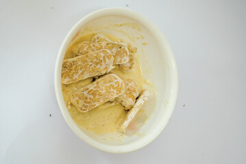 Raw tempeh that has been cut in a white bowl containing dough, with a white background.