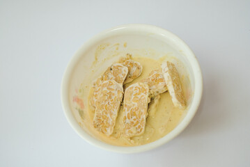 Raw tempeh that has been cut in a white bowl containing dough, with a white background.