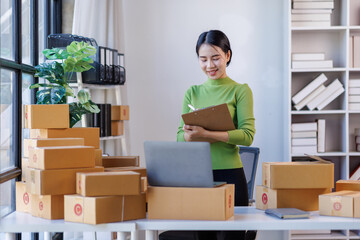 Portrait of Starting small businesses SME owners young female entrepreneurs check online orders on clipboard and laptop to prepare to pack the boxes, sell to customers, sme business ideas online.