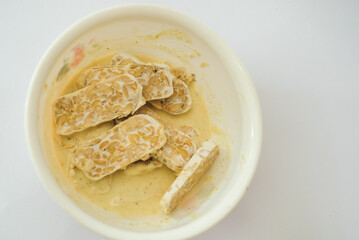 Raw tempeh that has been cut in a white bowl containing dough, with a white background.