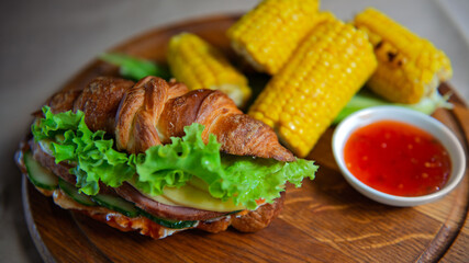 A gourmet croissant sandwich with fresh vegetables and ham, paired with corn on the cob and chili sauce, showcasing rustic culinary elegance.