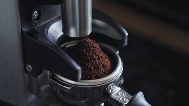 A handheld angle closeup shot of grounded coffee falling from a coffee grinder into a portafilter of a coffee machine