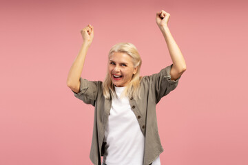 Excited mature, middle aged woman celebrating with raised arms on pink background