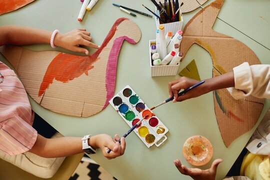 Top down view at group of children painting cardboard dinosaur figures using colorful watercolors during art and craft class copy space