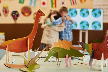 Close up background image of colorful handmade dinosaur figures painted during art and craft class with group of kids hugging in background copy space