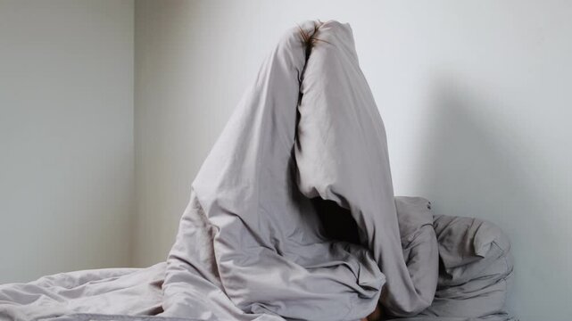 Young depressed woman hiding under a blanket in a gray, minimalist room, struggling with mental health issues, self-doubt, and emotional distress.