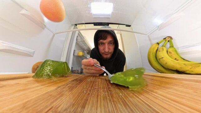 A close-up view of a man leaning into the refrigerator, finishing the remaining green jelly with a spoon. The scene highlights the glossy texture of the jelly alongside bananas and grapefruit.
