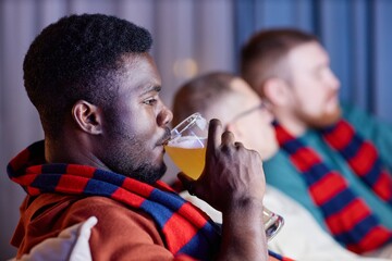 Side view closeup of adult African American man drinking beer from tall glass while sitting on couch and watching sports match on TV