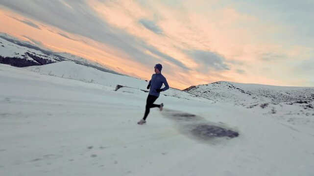Young Caucasian male athlete runner walking in pashmina and warm clothes in winter landscape in mountains surrounded by snow and mountains at sunset. Jogging at dusk. Aerial view from fpv sports drone