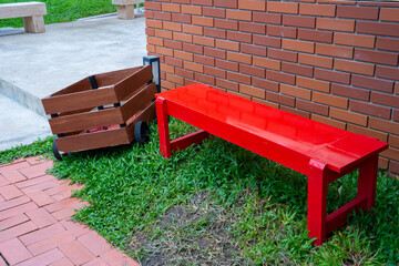 A vibrant red bench beside a wooden cart on green grass, against a brick wall, creating an inviting...
