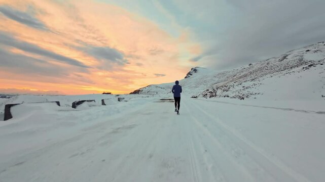 Young Caucasian male athlete runner walking in pashmina and warm clothes in winter landscape in mountains surrounded by snow and mountains at sunset. Jogging at dusk. Aerial view from fpv sports drone