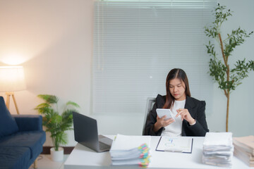 Young businesswoman calculating and reviewing financial documents while using a calculator and laptop, surrounded by stacks of paperwork at her organized office desk