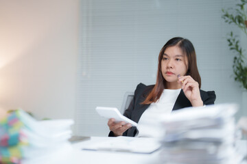 Businesswoman is calculating and thinking about her business while using a calculator and reviewing documents at her desk in a bright office