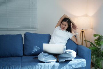 Young Asian woman is sitting on comfortable sofa and stretching her arms while working on laptop at home, taking a break from work to relax muscles