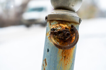 Close-up of a weathered, rusted metal pole with an attached rusty metal loop, covered in streaks of corrosion and decay. The snow-covered ground and soft-focus background suggest a wintry environment