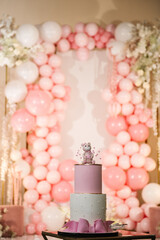 Cake is decorated with bear figure, stars and a big bow for baby girl for 1, 2, 3 years. Delicious reception at birthday party. Arch decorated with pink, white balloons. Trendy baptism cake for girl.
