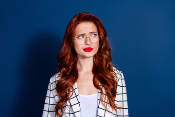 Charming young woman with red hair and glasses in a checkered suit stands against a dark blue...