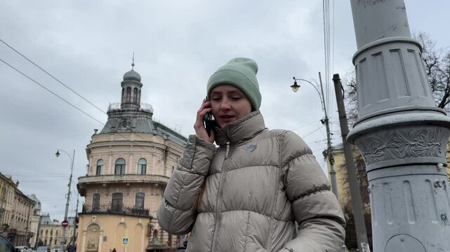 Young woman wearing winter clothes chatting on her phone near a lamppost in ukraine, on a cloudy day