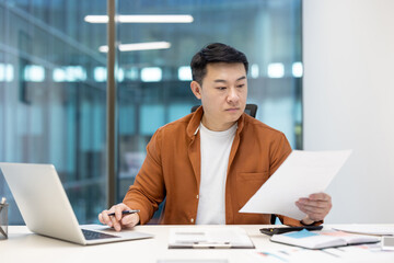 Asian man mature businessman in an office setting reviewing paperwork while working on a laptop, representing productivity, professionalism, and focus in a corporate environment.