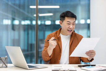Asian man expressing excitement over positive outcomes while working at a modern office desk. The...
