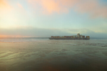 Hazy winter landscape at dawn, with cargo boat full of containers seen from the coast moving forward on the St. Lawrence river in the fog, Sainte-P&eacute;tronille, Island of Orleans, Quebec, Canada