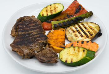 Carne a la parrilla con verduras en un plato blanco sobre un fondo blanco.