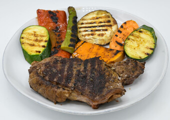 Carne a la parrilla con verduras en un plato blanco sobre un fondo blanco.