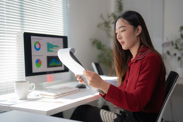 Young asian businesswoman reviewing financial reports and data analysis on her computer while holding documents, working diligently in a modern office setting