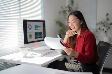 Young professional reviewing paperwork while using a computer for data visualization, smiling and holding a pen, fully engaged in business analysis and strategy development