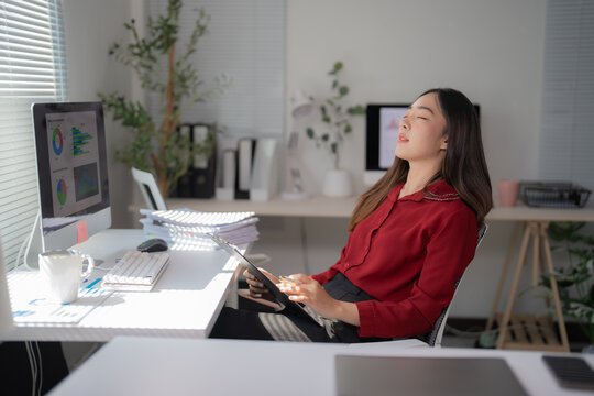 Asian businesswoman relaxing with eyes closed while holding a clipboard and sitting at her desk in a modern office, enjoying a break after analyzing financial data