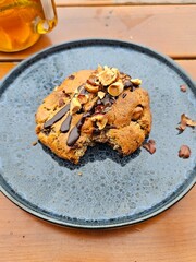 Un cookie fait maison garni de pépites de chocolat, de noisettes croquantes et d’un filet de chocolat fondant repose sur une assiette bleue texturée, posée sur une table en bois.