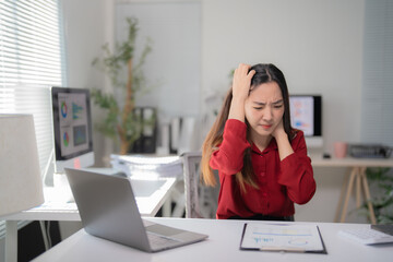 Young business woman suffering from strong headache after hard working day sitting at her workplace with laptop and documents in modern office, feeling tired exhausted and painful