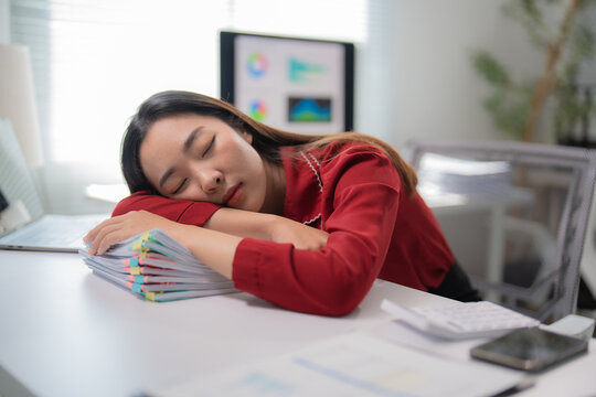 Asian businesswoman in red blouse is sleeping on a pile of documents at her desk, overwhelmed by work and fatigue in a modern office setting - Powered by Adobe