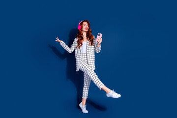 Young woman with red hair in stylish checkered suit, dancing joyfully against dark blue background, holding smartphone, wearing headphones